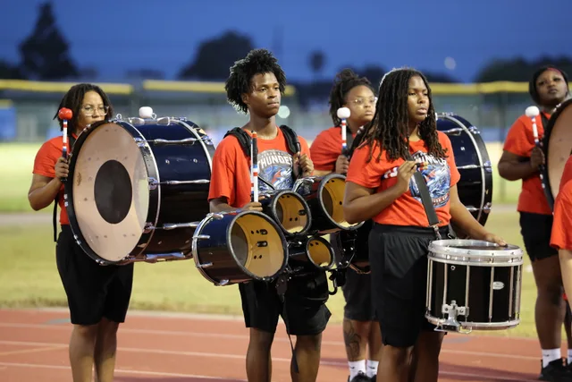 The drumline assembled beside the track at dusk, in red shirts beneath a blue evening sky.
